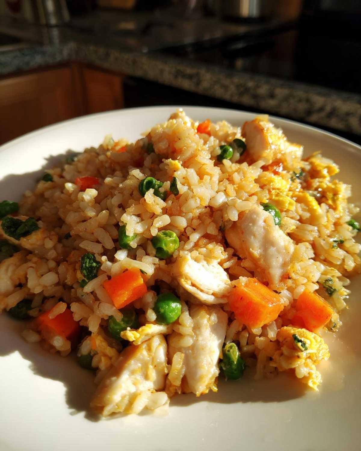 A close-up photo of a serving of amazing chicken fried rice with visible chunks of chicken, carrots, and peas on a white plate.