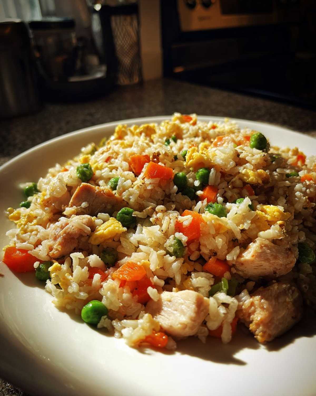 A close-up, sunlit view of a plate piled high with amazing chicken fried rice, featuring white rice, diced chicken, carrots, peas, and scrambled egg.