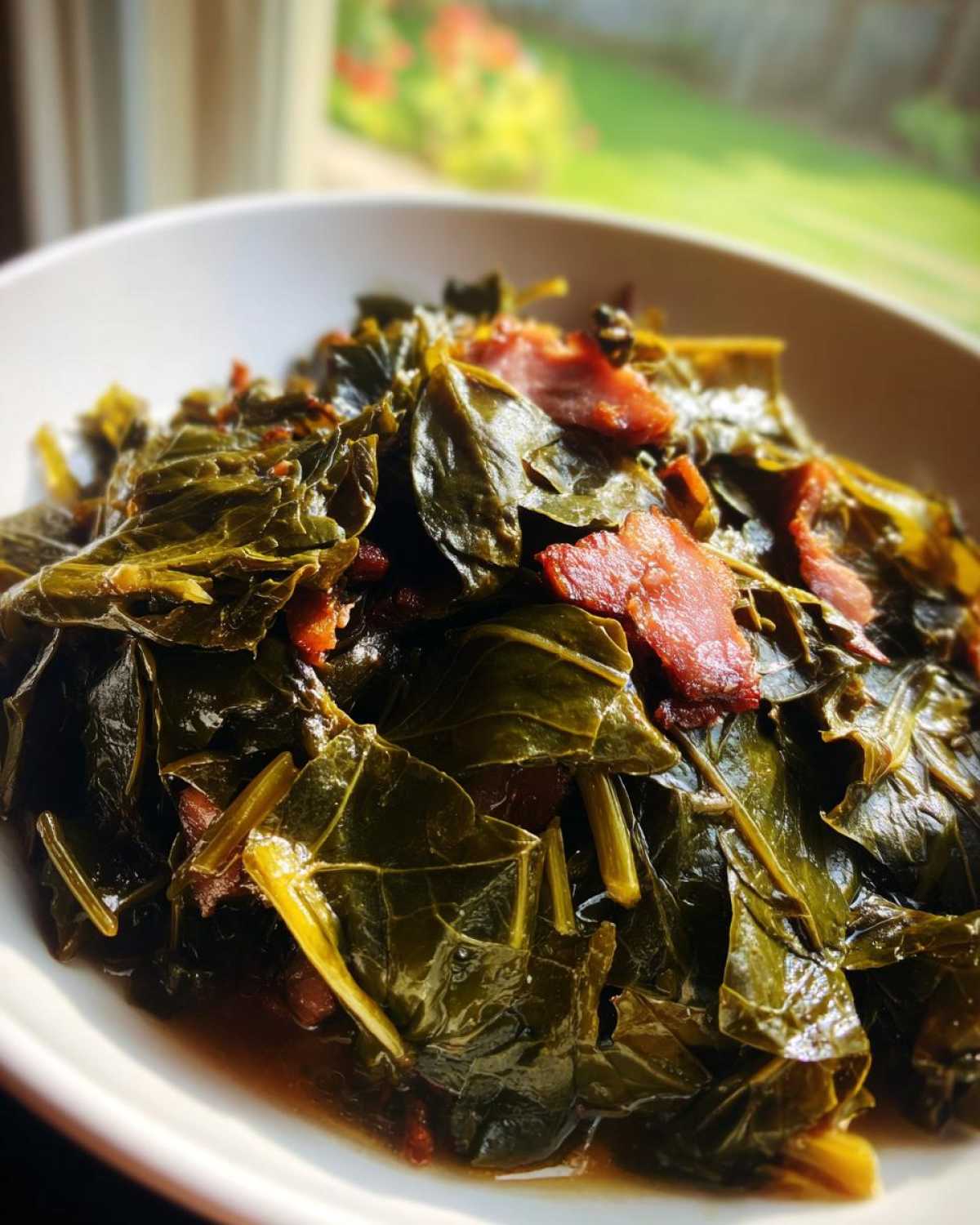 Close-up of tender, slow-cooked collard greens served in a white bowl with pieces of smoked meat.