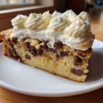 A close-up of a slice of amazing cookie cake recipe, featuring chocolate chips and piped whipped cream frosting.