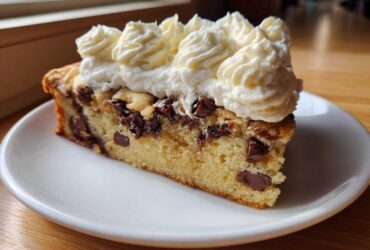 A close-up of a slice of amazing cookie cake recipe, featuring chocolate chips and piped whipped cream frosting.