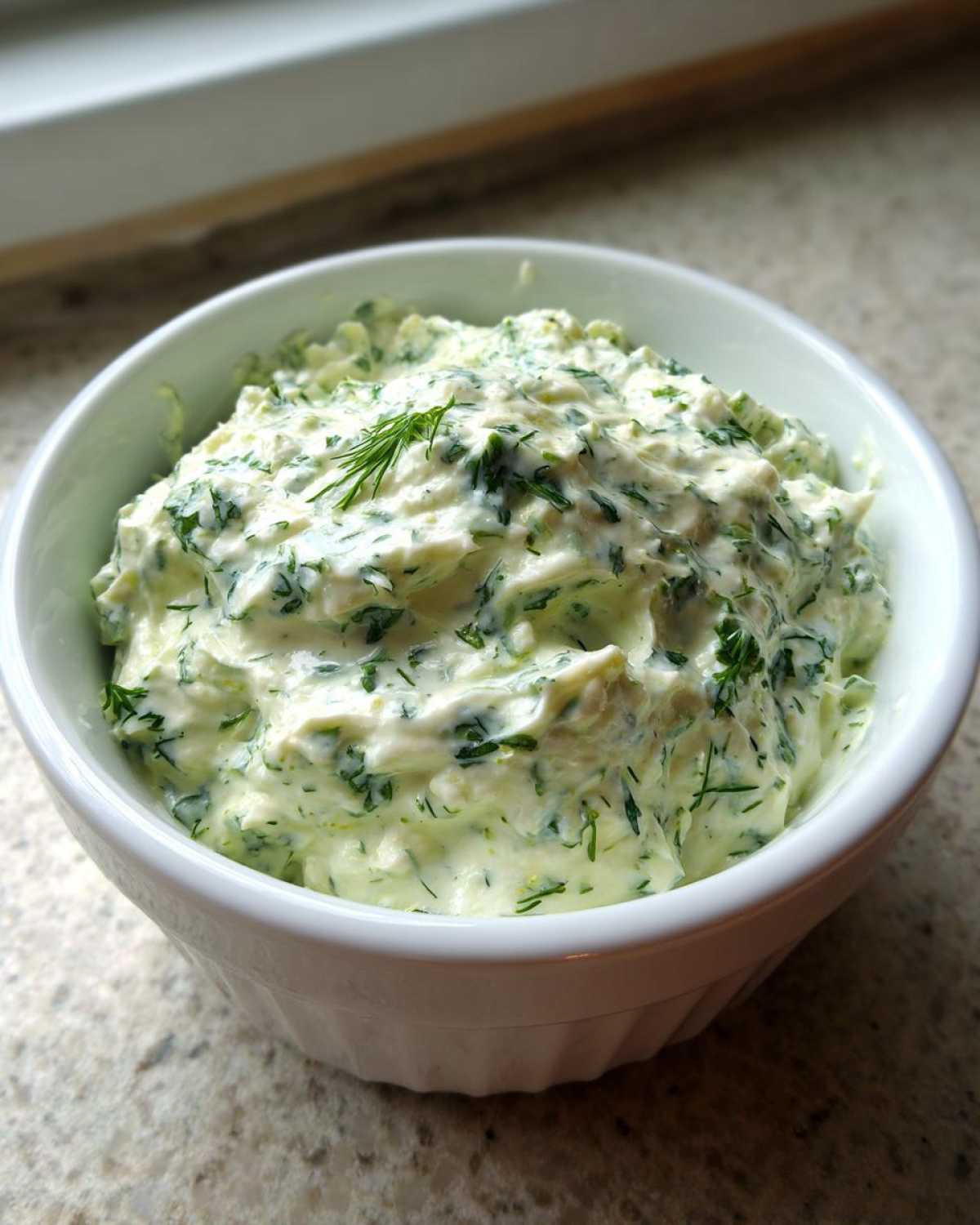 Close-up of creamy, bright green dill dip generously mixed with fresh herbs in a small white bowl.