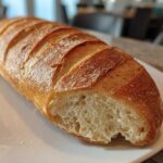 A golden-brown, freshly baked loaf of French bread with a crisp, scored crust, cut open to show the soft interior.