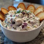 A creamy, chunky hoagie dip garnished with herbs and served in a white bowl surrounded by toasted baguette slices.