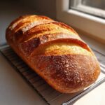A golden-brown, crusty loaf of homemade Italian bread cooling on a wire rack near a window.