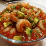 Close-up of a glass bowl filled with amazing mexican shrimp cocktail, featuring plump shrimp, avocado chunks, and a rich tomato-based sauce.