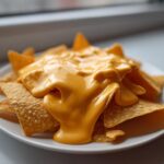 Close-up of tortilla chips smothered in thick, smooth nacho cheese sauce on a white plate.