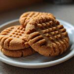 Four delicious peanut butter cookies stacked on a small white plate, showing the classic criss-cross fork marks.