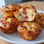 A stack of golden brown pizza muffins with melted, bubbly cheese topping, one is broken open showing pepperoni.