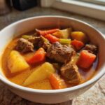 A close-up of a white bowl filled with rich, orange-hued pork stew featuring tender meat chunks, potatoes, and carrots.