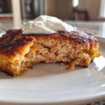 Close-up of a perfectly seared salmon cake with a bite taken out, showing flaky interior and topped with a dollop of white sauce.