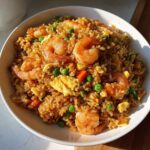 A close-up overhead shot of a white bowl filled with savory shrimp fried rice, featuring large shrimp, green peas, carrots, and scrambled egg.