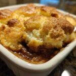 A close-up view of a scoop being taken out of a warm, bubbling apple cobbler with a golden, biscuit-like topping.