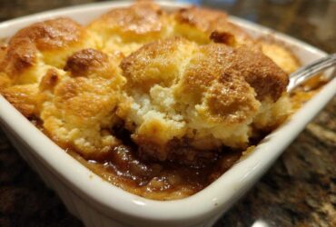 A close-up view of a scoop being taken out of a warm, bubbling apple cobbler with a golden, biscuit-like topping.