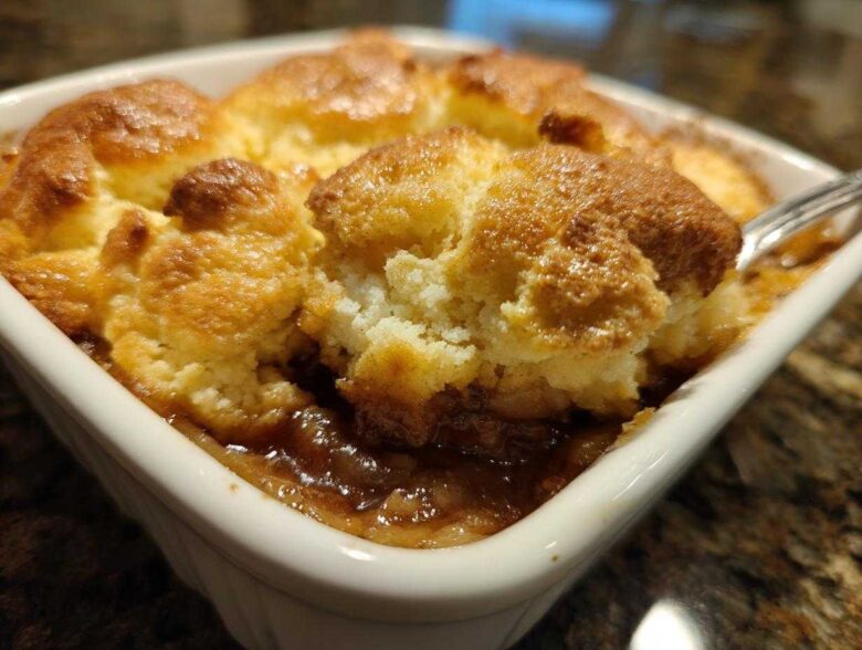 A close-up view of a scoop being taken out of a warm, bubbling apple cobbler with a golden, biscuit-like topping.