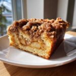 A close-up of a moist slice of apple crumb cake with visible apple chunks and a thick cinnamon streusel topping.