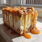 A thick slice of apple pie cheesecake dripping with rich caramel sauce, set against a bright window.