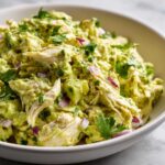 A close-up shot of creamy avocado chicken salad mixed with chunks of avocado, red onion, and cilantro in a white bowl.
