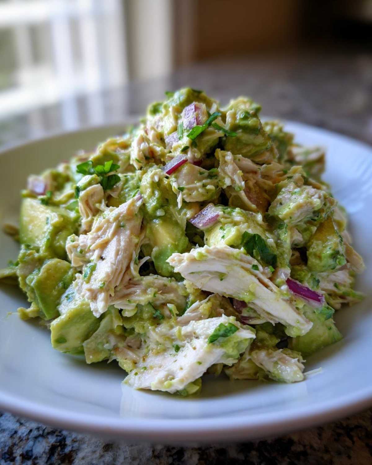 A close-up view of creamy avocado chicken salad featuring shredded chicken, chunks of avocado, and red onion.