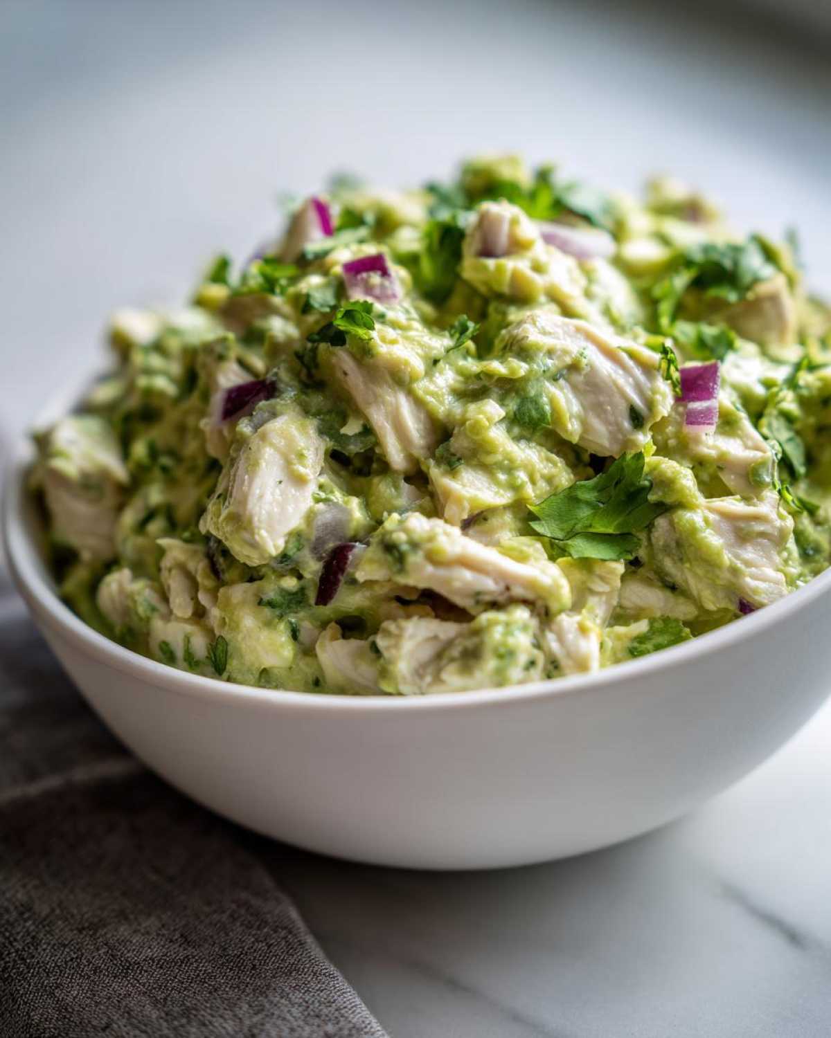 Close-up of creamy avocado chicken salad mixed with red onion and cilantro in a white bowl.