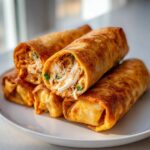 Close-up of four golden-brown baked Chimichangas stacked on a white plate, showing the shredded chicken filling inside.