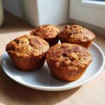 Four freshly baked banana protein muffins with a cinnamon-sugar topping resting on a white plate.