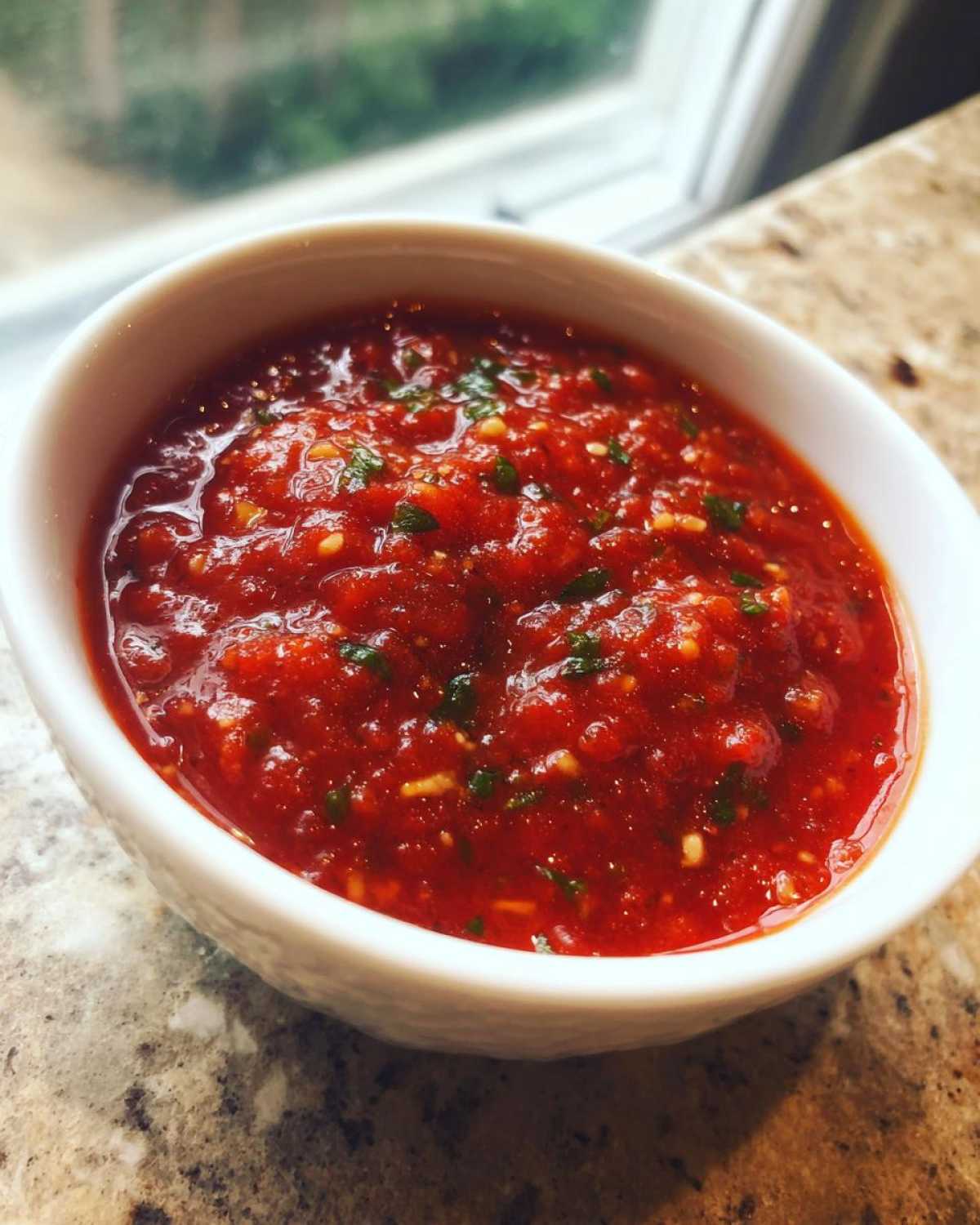 A close-up of vibrant, chunky homemade pizza sauce with visible herbs and garlic in a white bowl.