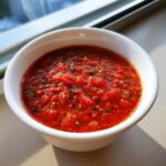 A close-up of vibrant, chunky pizza sauce seasoned with herbs, served in a small white bowl near a window.