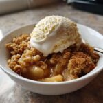 A warm bowl of homemade apple crumble topped with a melting scoop of vanilla ice cream.