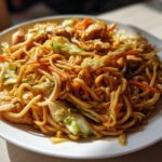 A close-up of a hearty plate of chicken chow mein recipe, featuring noodles, chicken pieces, cabbage, and carrots.