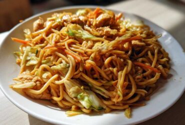 A close-up of a hearty plate of chicken chow mein recipe, featuring noodles, chicken pieces, cabbage, and carrots.