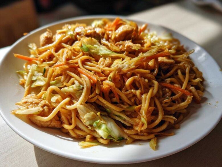 A close-up of a hearty plate of chicken chow mein recipe, featuring noodles, chicken pieces, cabbage, and carrots.