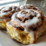 Two freshly baked, gooey cinnamon roll pastries covered in thick white icing, sitting on a white plate.