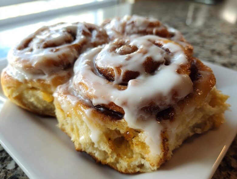 Two freshly baked, gooey cinnamon roll pastries covered in thick white icing, sitting on a white plate.