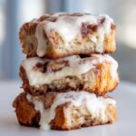 Close-up of three fluffy, iced cinnamon rolls stacked vertically, dripping with white cream cheese frosting.