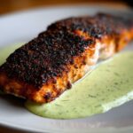 Close-up of a perfectly cooked blackened salmon fillet resting on a pool of light green dill sauce.