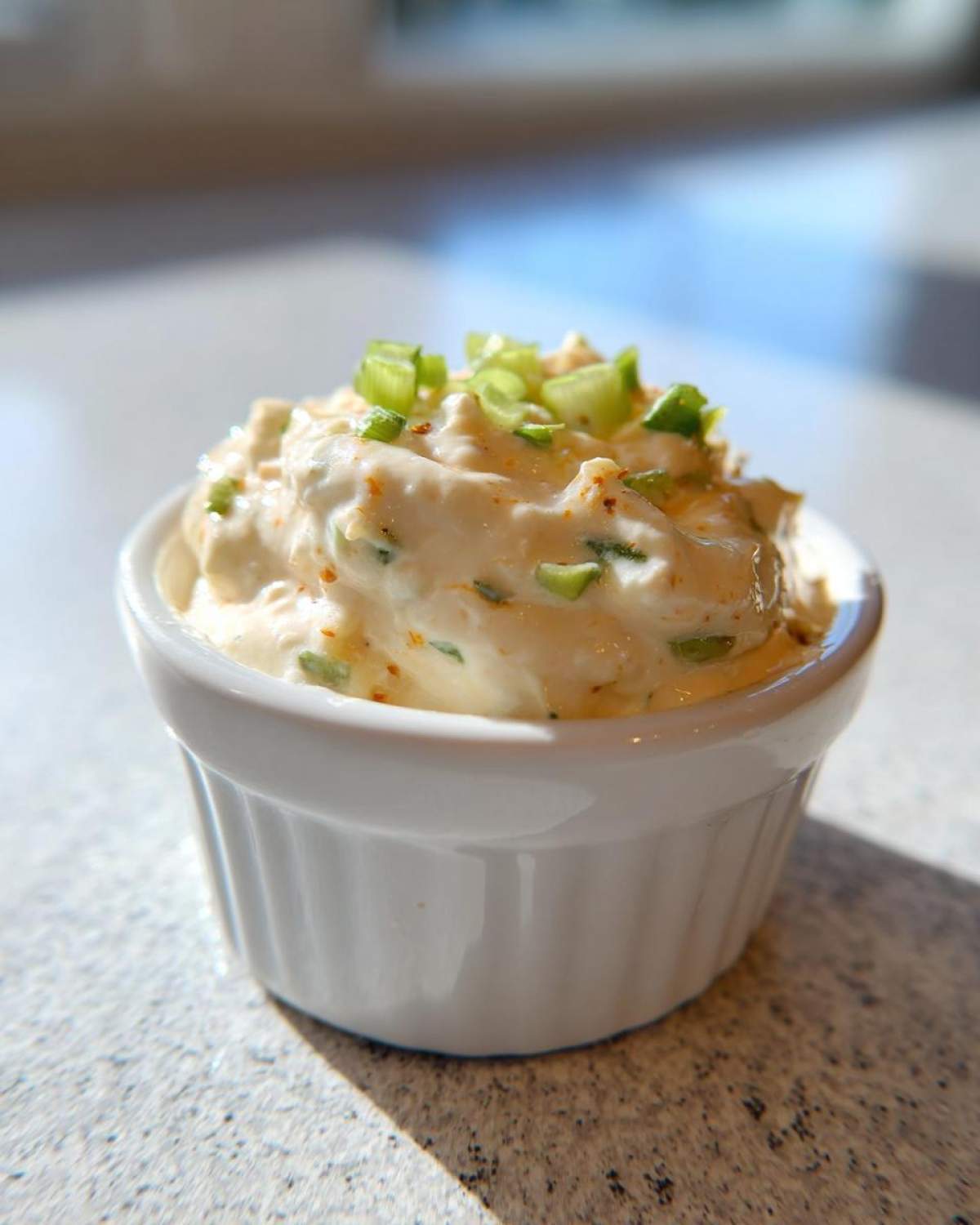 Close-up of a small white ramekin filled with thick, creamy remoulade sauce, topped with chopped green onions.