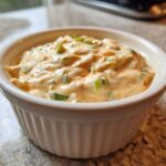 A close-up of a small white ramekin filled with creamy, orange-tinted remoulade sauce topped with chopped green onions.