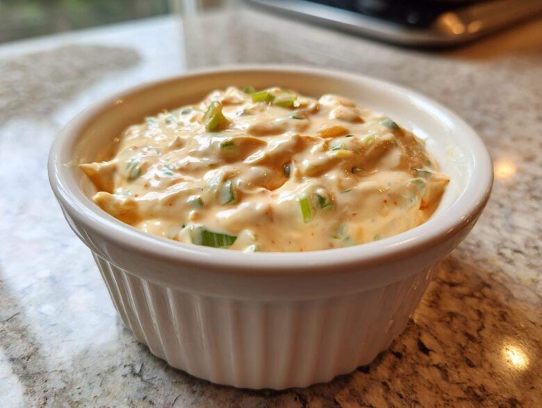 A close-up of a small white ramekin filled with creamy, orange-tinted remoulade sauce topped with chopped green onions.