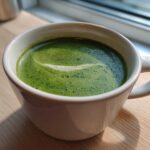 A close-up of bright green zucchini soup with a swirl of foam in a white mug, suggesting a healthy, low-calorie option.