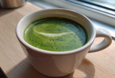 A close-up of bright green zucchini soup with a swirl of foam in a white mug, suggesting a healthy, low-calorie option.