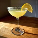 A bright yellow lemon drop cocktail served in a coupe glass with a sugared rim and lemon slice garnish.