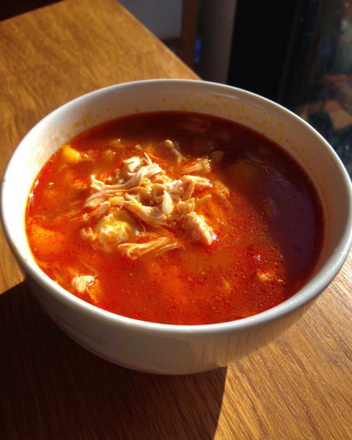 A white bowl filled with vibrant red chicken pozole soup, topped with shredded chicken, sitting on a wooden table.