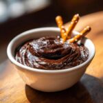 A close-up of rich, dark chocolate dip swirled in a white bowl, garnished with pretzel sticks.