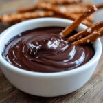 Close-up of smooth, glossy chocolate dip in a white bowl with pretzel sticks ready for dipping.