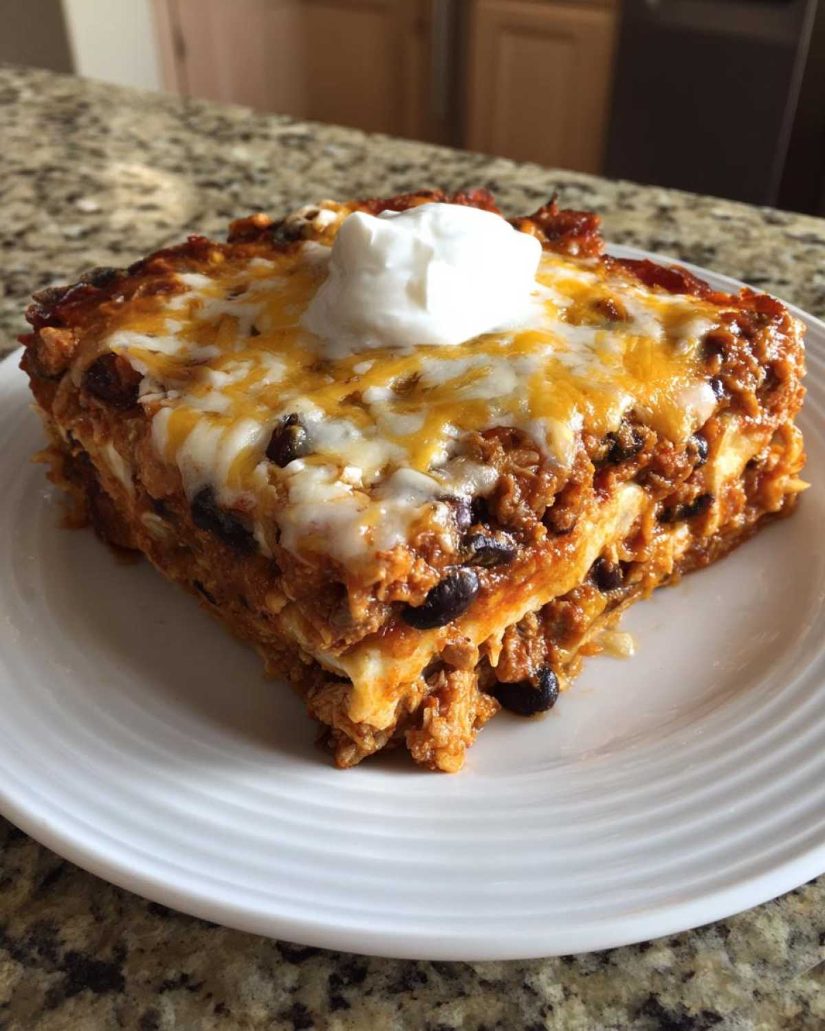 A square slice of cheesy, layered burrito bake topped with melted cheese and a dollop of sour cream.