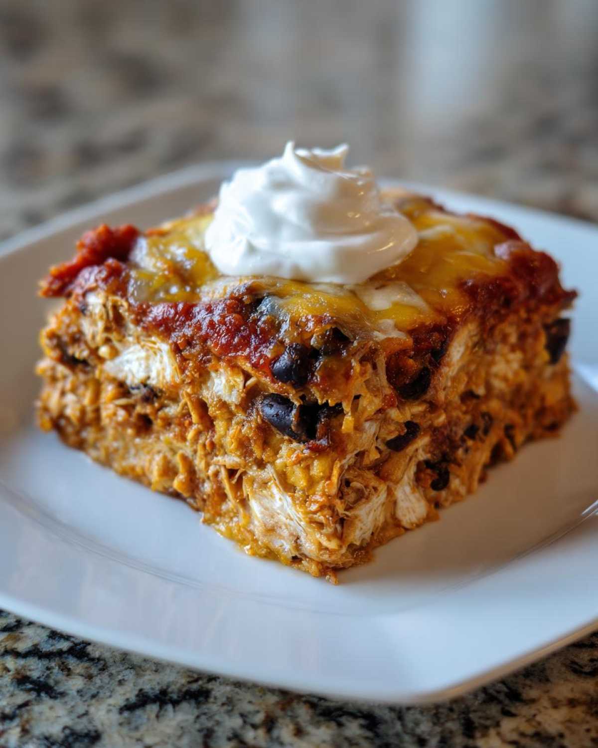 A square slice of cheesy, layered burrito bake topped with salsa and sour cream on a white plate.