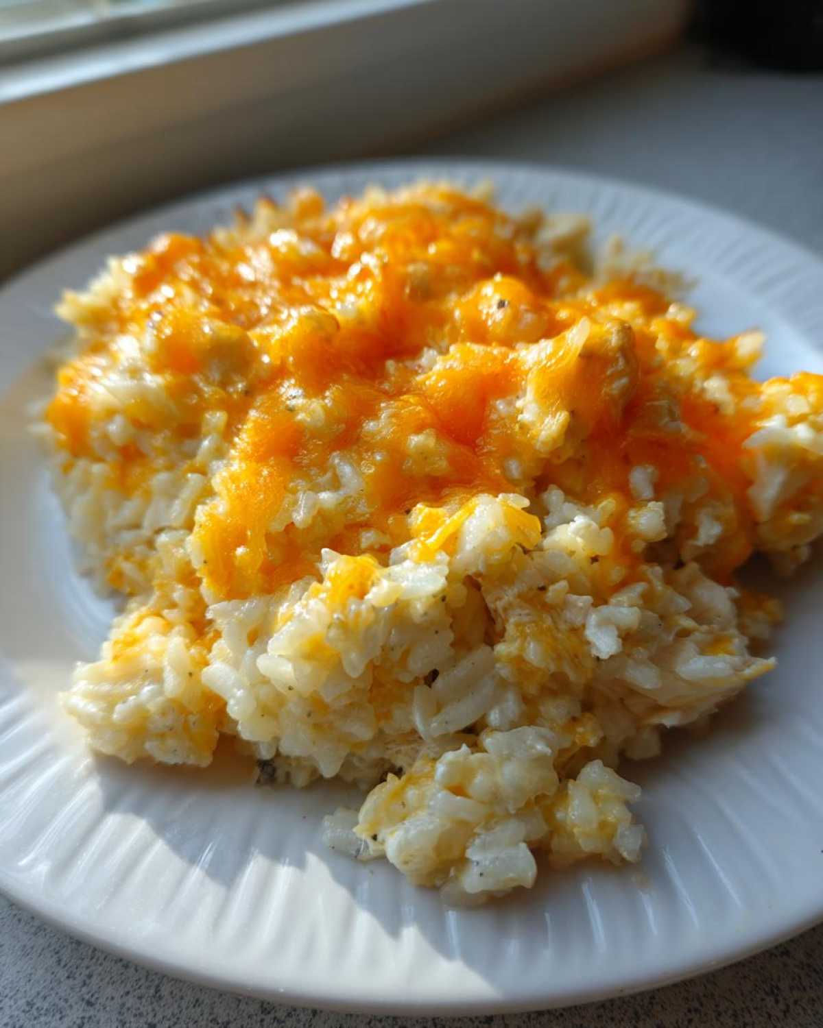 Close-up of cheesy chicken and rice topped with melted bright orange cheddar cheese on a white plate.