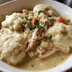 A close-up of a white bowl filled with creamy chicken and dumplings, topped with fresh parsley.