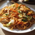 Close-up of a plate piled high with saucy chicken chow mein recipe, featuring noodles, chicken pieces, carrots, and cabbage.